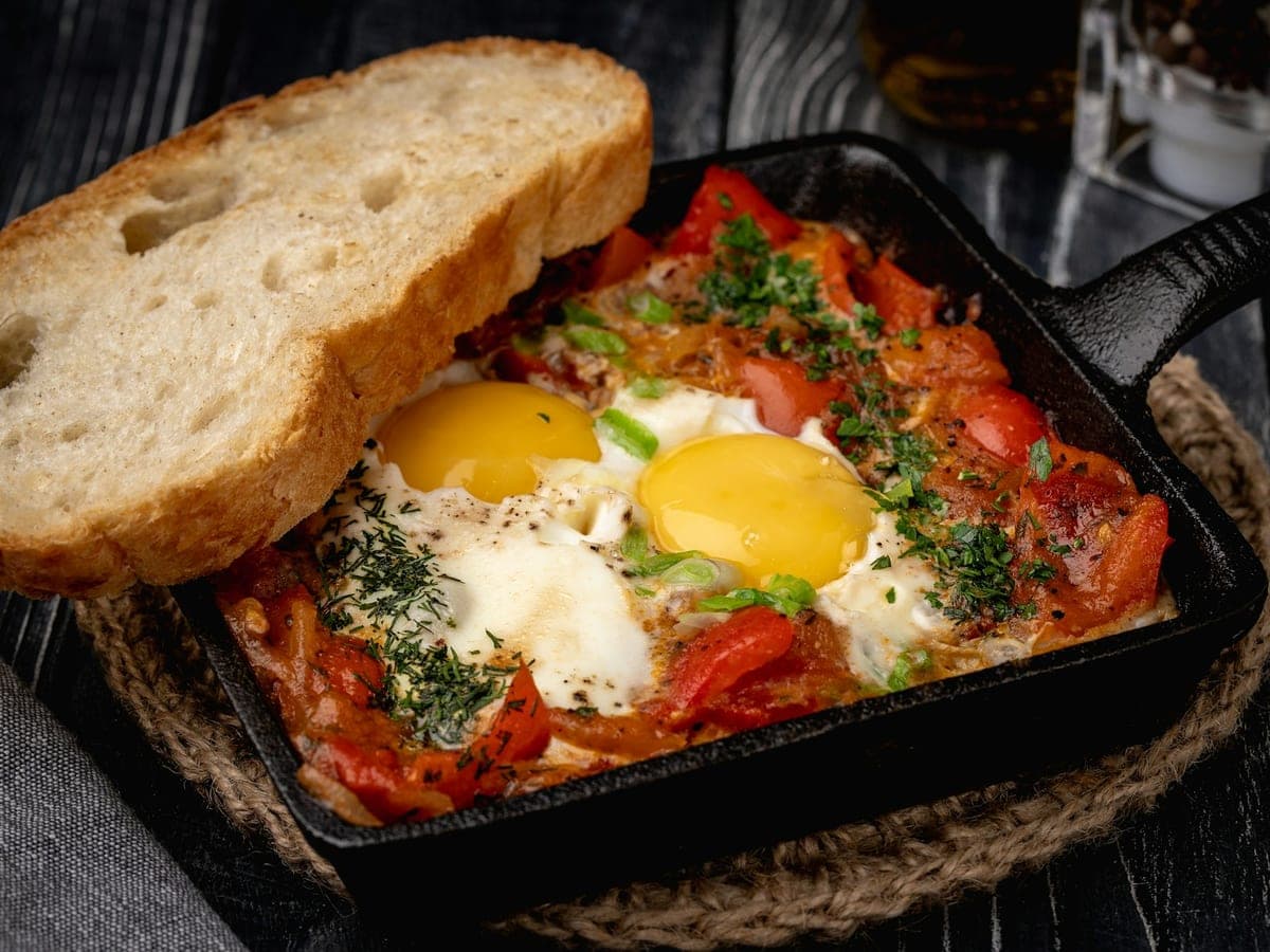 Shakshuka in een metalen pan, eieren gepocheerd in pittige tomatensaus, met brood en salade ernaast op tafel.