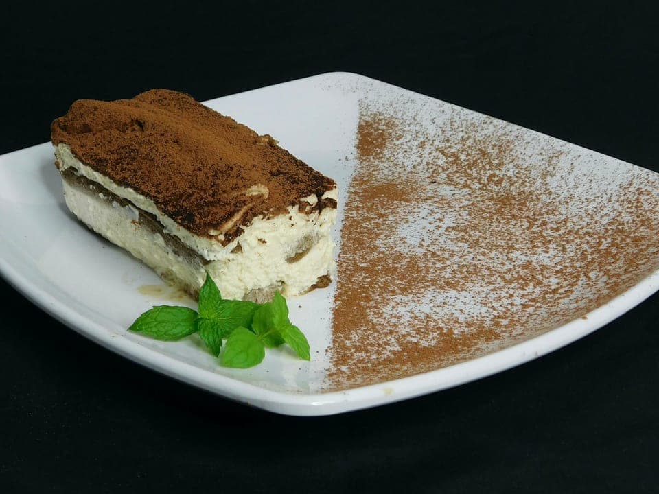 Een vierkant stuk tiramisu op een wit bord, met een dikke laag cacaopoeder bovenop.
